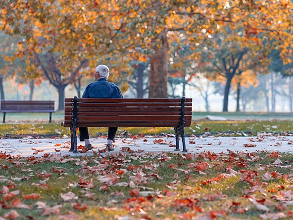 Older man sits alone on park bench surrounded by autumn leaves